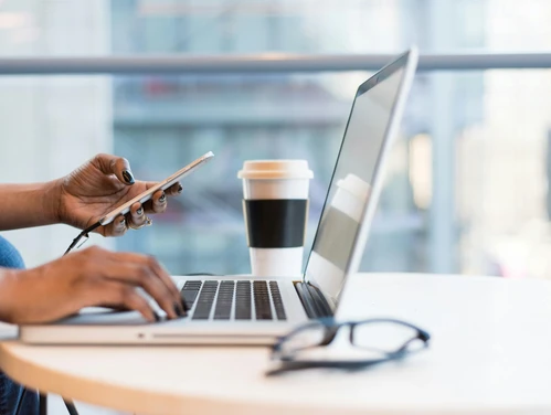 A hand holds a smartphone, while the other hovers over the keyboard of an open laptop