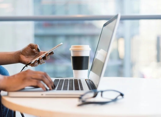 A hand holds a smartphone, while the other hovers over the keyboard of an open laptop